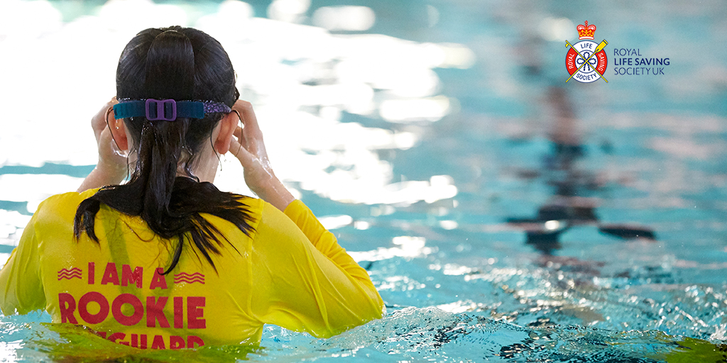 Child wearing a yellow shirt with red writing reading "Rookie Lifeguard" standing in a pool and putting on swimming goggles, seen from behind.